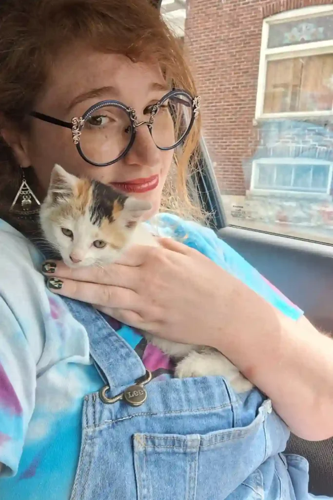 A smiling woman with glasses and curly hair holds a small calico kitten close to her chest while sitting in a car.